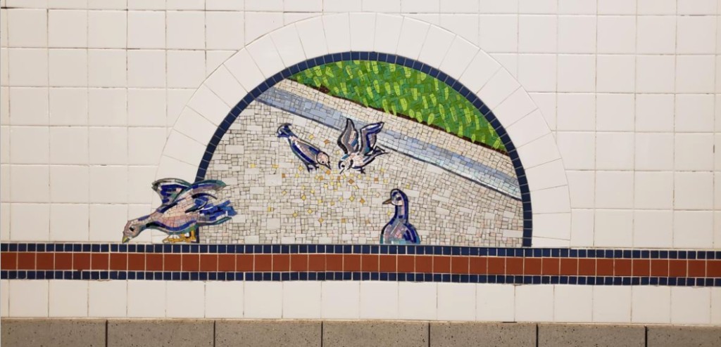 A half-moon mosaic from Broadway Diary at the 8th Street–NYU station, showing pigeons pecking at crumbs on a pavement beside a strip of green, set into white subway tile