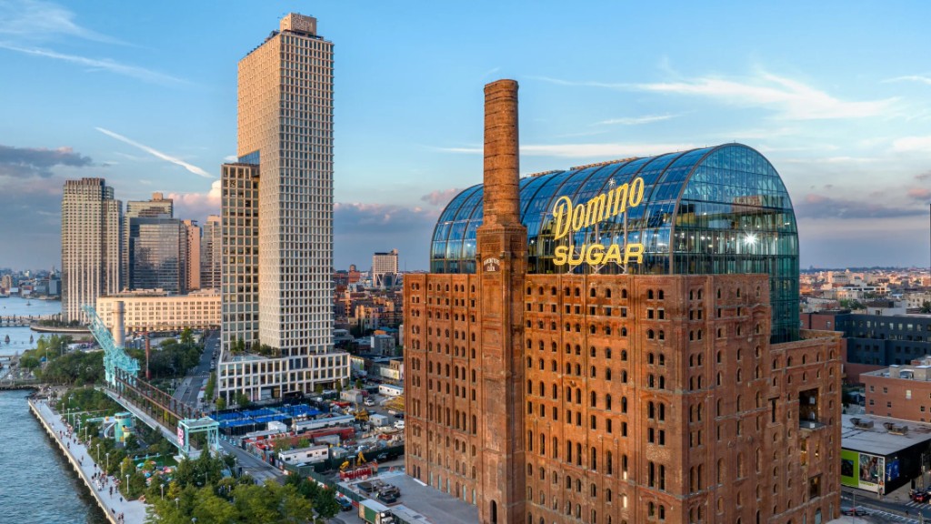 The restored Domino Sugar Refinery at sunset, its brick facade and chimney topped by a vaulted glass roof bearing the yellow Domino Sugar sign, beside the East River waterfront in Williamsburg