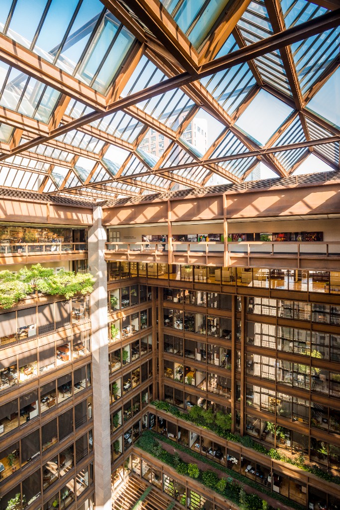 Looking up through the Ford Foundation Atrium to its geometric glass ceiling, framed by weathered steel beams and tiers of glass-fronted offices with planted balconies