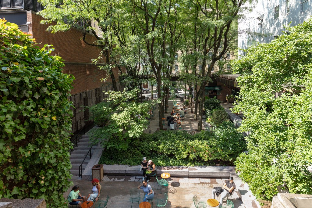 Greenacre Park from above: people at small tables on a stone terrace beneath a canopy of honey locust trees, with steps leading up to a raised seating area