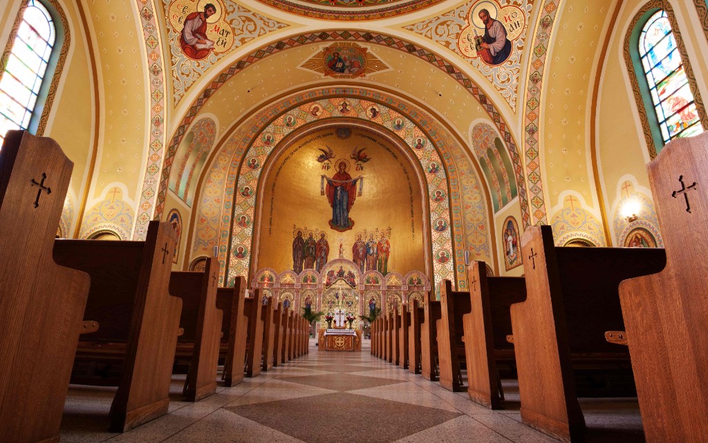The interior of St. George Ukrainian Catholic Church, looking down the aisle between wooden pews toward a gilded apse painted with icons