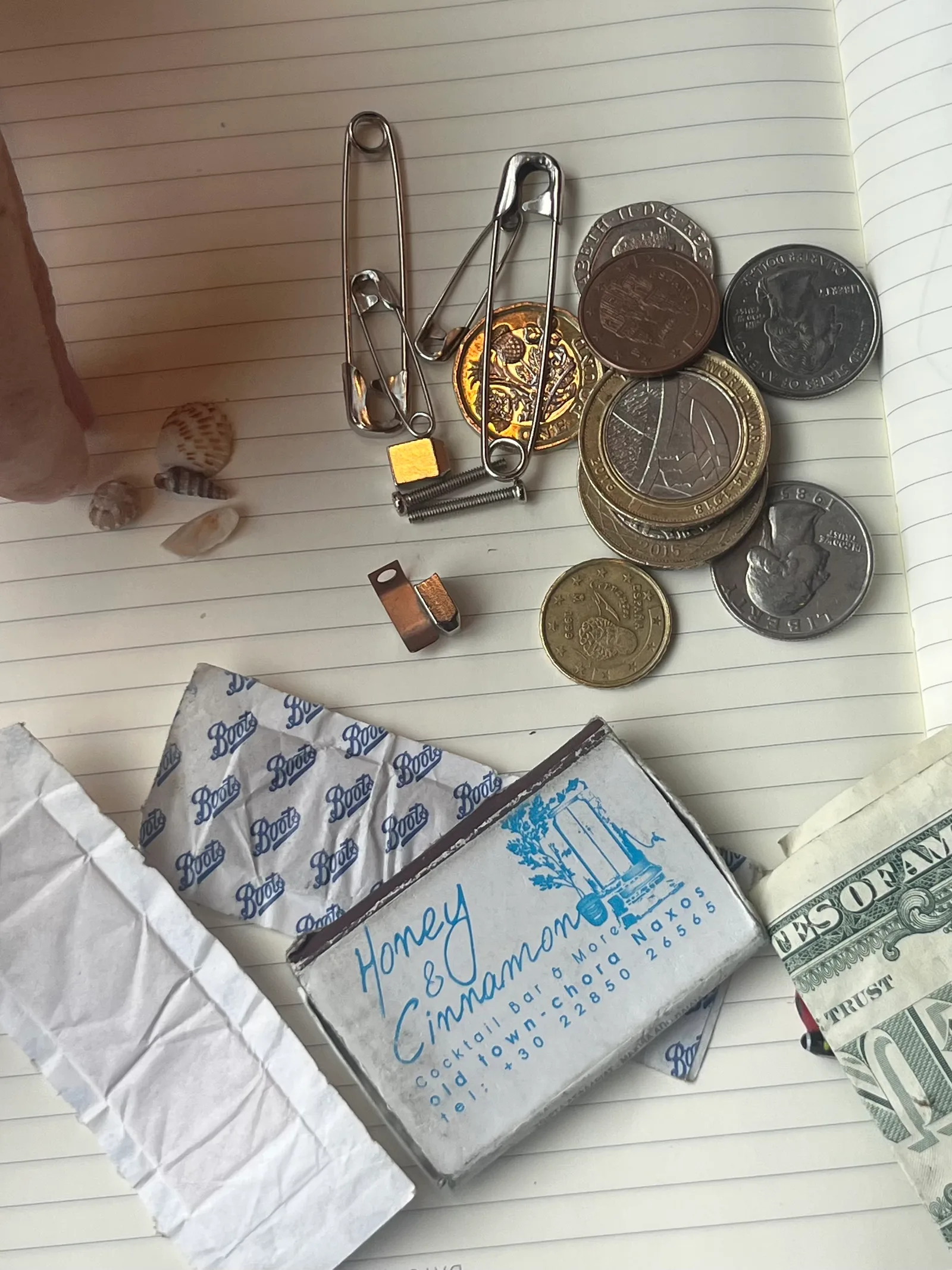 Travel keepsakes on a lined notebook page: pound, euro, and US coins, safety pins, small shells, a Boots wrapper, a dollar bill, and a matchbox from a cocktail bar in Naxos