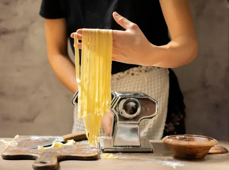 Person holding freshly cut pasta strips next to a pasta machine on a wooden table