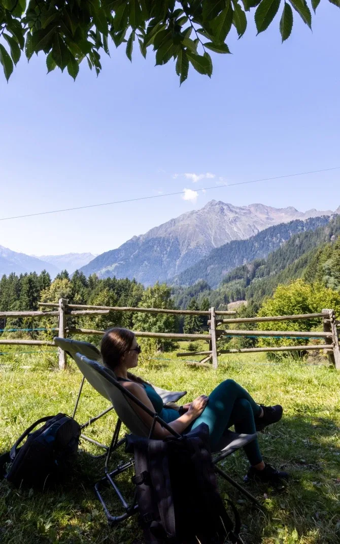 Person resting on a folding chair under tree shade with mountains and forest beyond a wooden fence