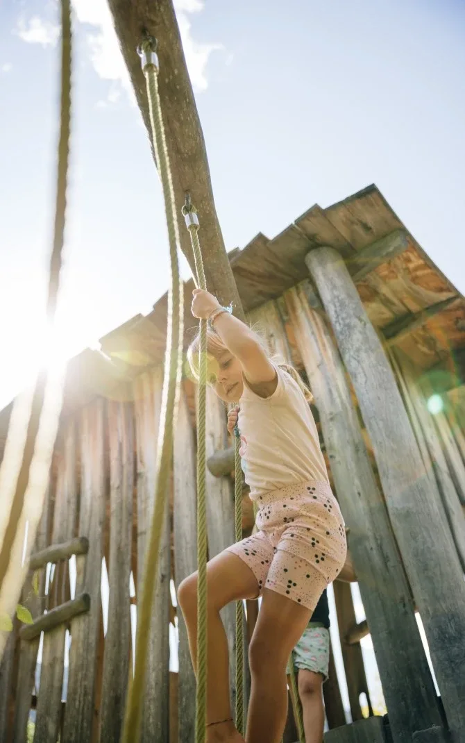 Child climbing a rope ladder on a wooden playground structure under sunlight
