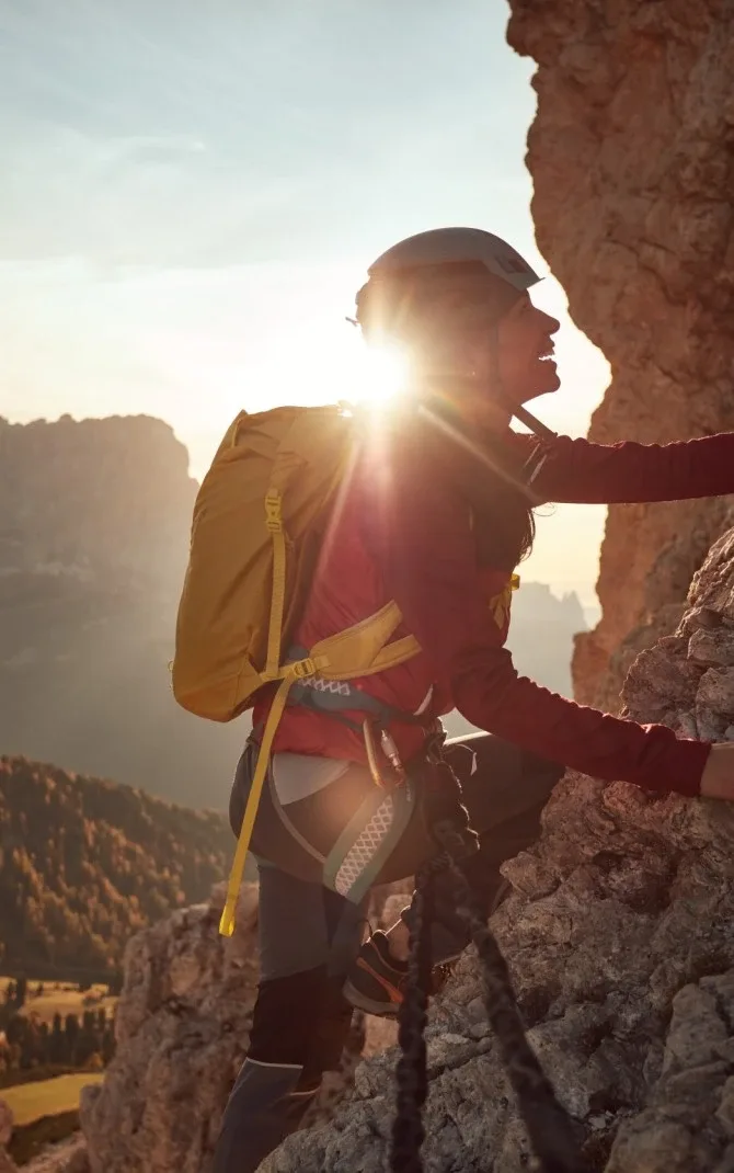 Person wearing climbing gear ascending a rocky cliff with a mountainous landscape in the background