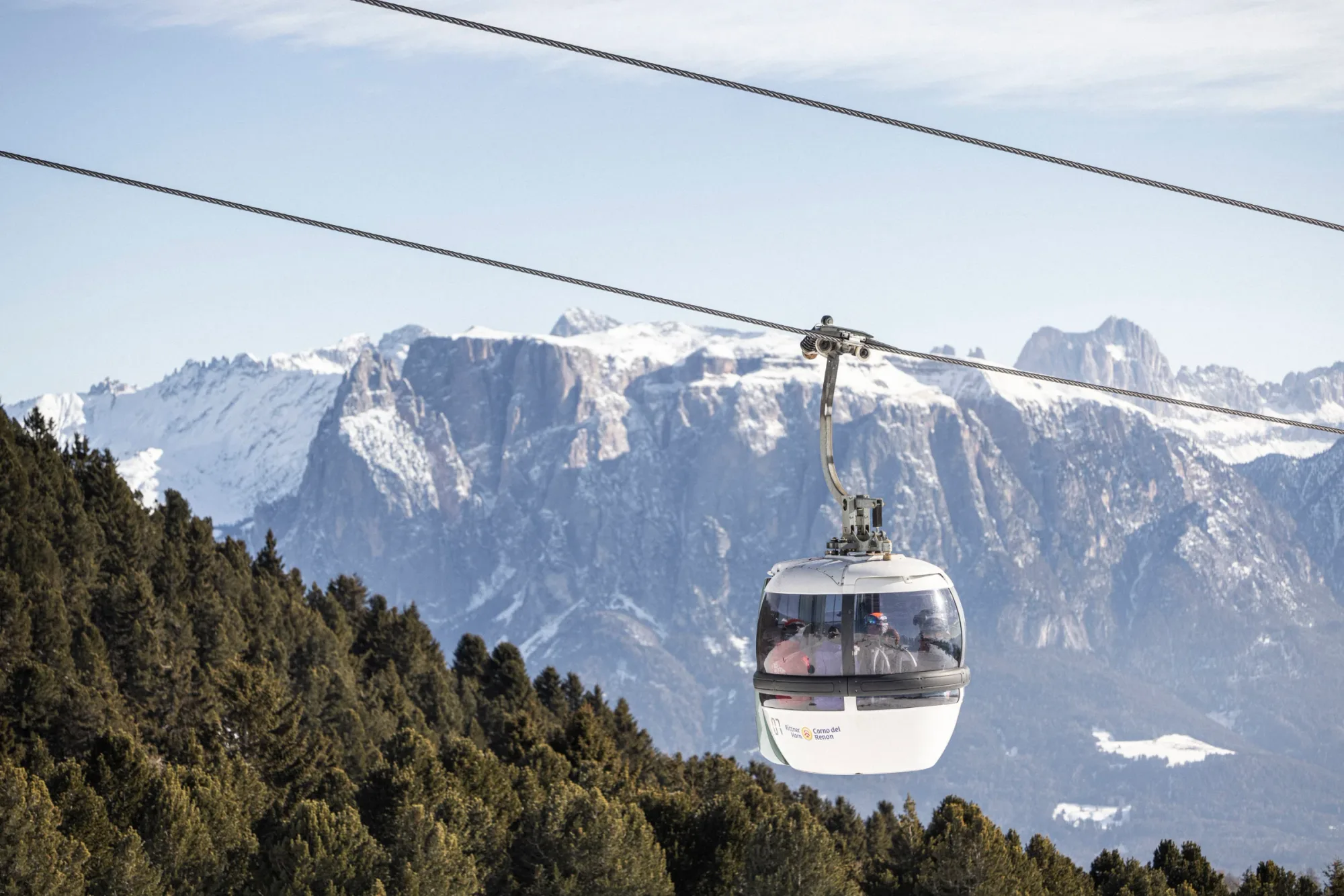 Cable car gondola carrying skiers above pine trees with snow-covered mountains in the background