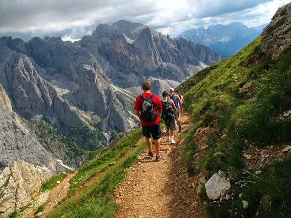Group hiking on a narrow mountain trail surrounded by rocky peaks and grassy slopes