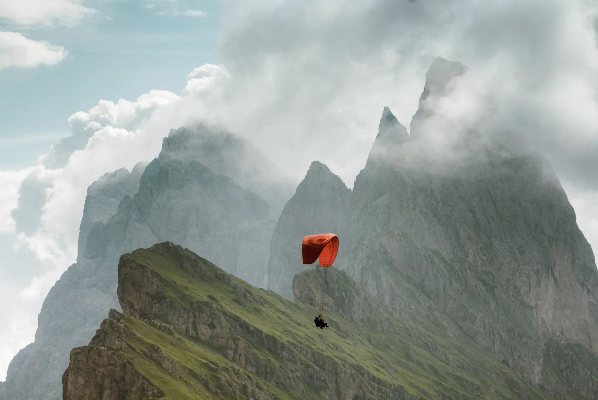Paraglider with red canopy flying near a green mountain ridge and misty rocky peaks
